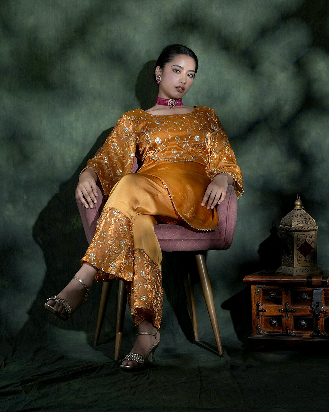A woman seated in a pink upholstered chair wearing an ornate golden embroidered traditional outfit with matching pants, styled with metallic heels and jewelry against a dark green textured backdrop. A small wooden cabinet with a lantern sits to the right.