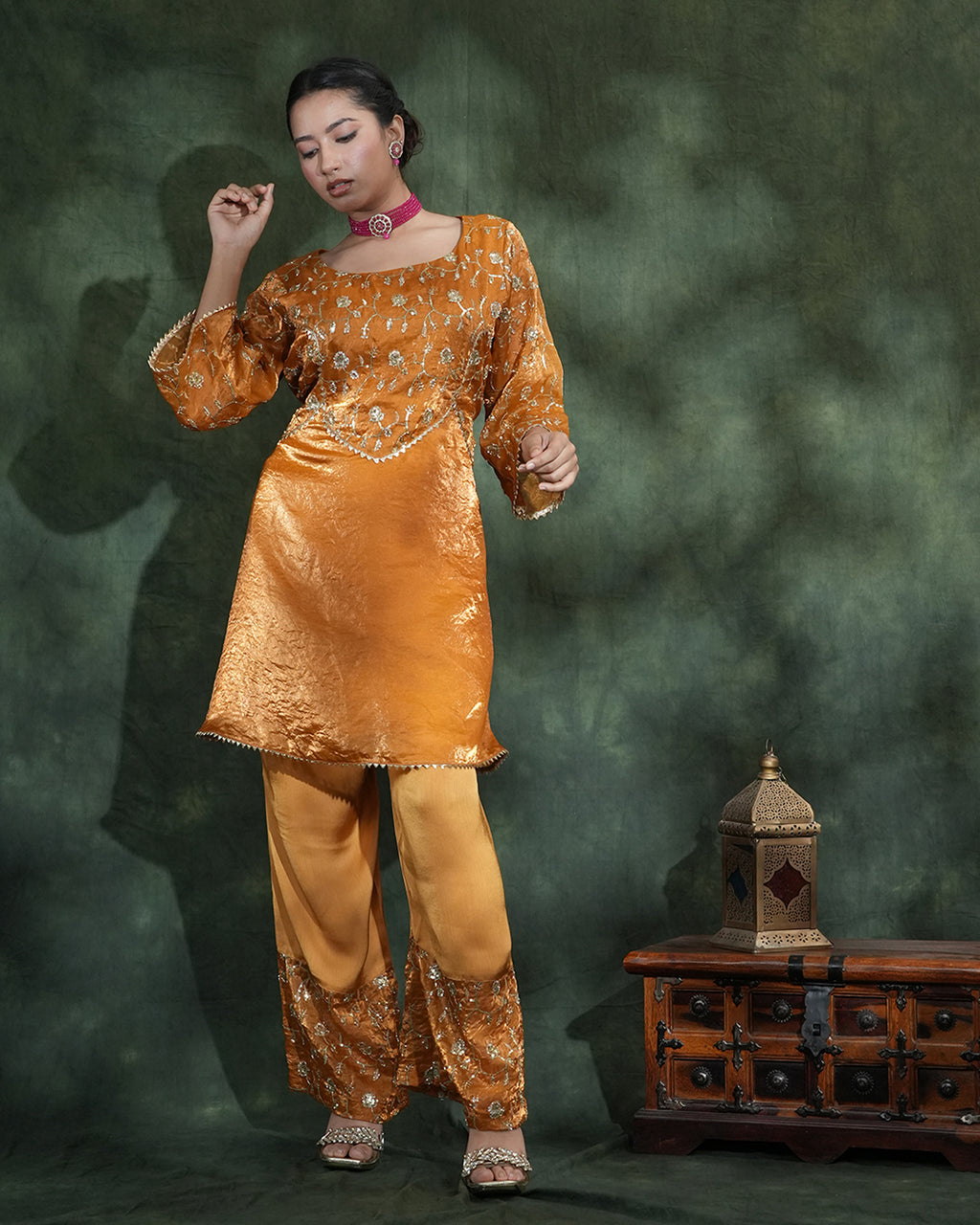 A woman models a traditional Indian outfit consisting of a golden-yellow kurta and trouser set with intricate silver embroidery, set against a dark green textured backdrop with a wooden chest.