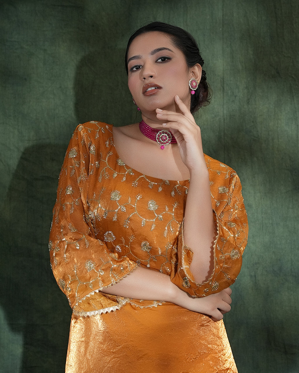 A woman modeling a traditional Indian outfit in a warm orange-gold color with intricate silver floral embroidery, paired with a matching skirt. She is posing against a dark textured green backdrop.