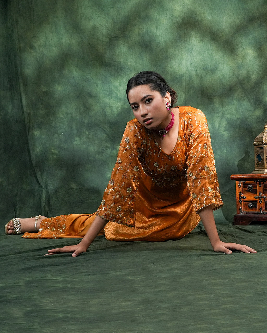 A woman in a metallic orange embroidered kurta and matching pants poses on a textured green backdrop, with a small wooden cabinet visible on the right side.