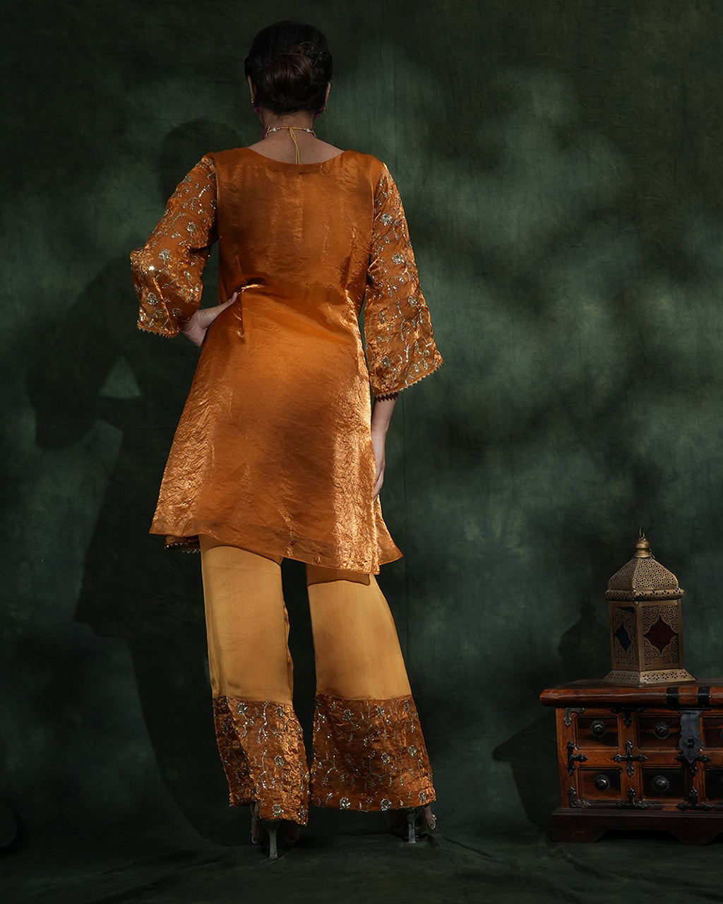 A woman stands with her back to the camera wearing a metallic mustard-gold tunic with embroidered sleeves and wide-leg trousers with matching embroidered hems, posed against a dark green textured backdrop with a small wooden cabinet and lantern at the side.