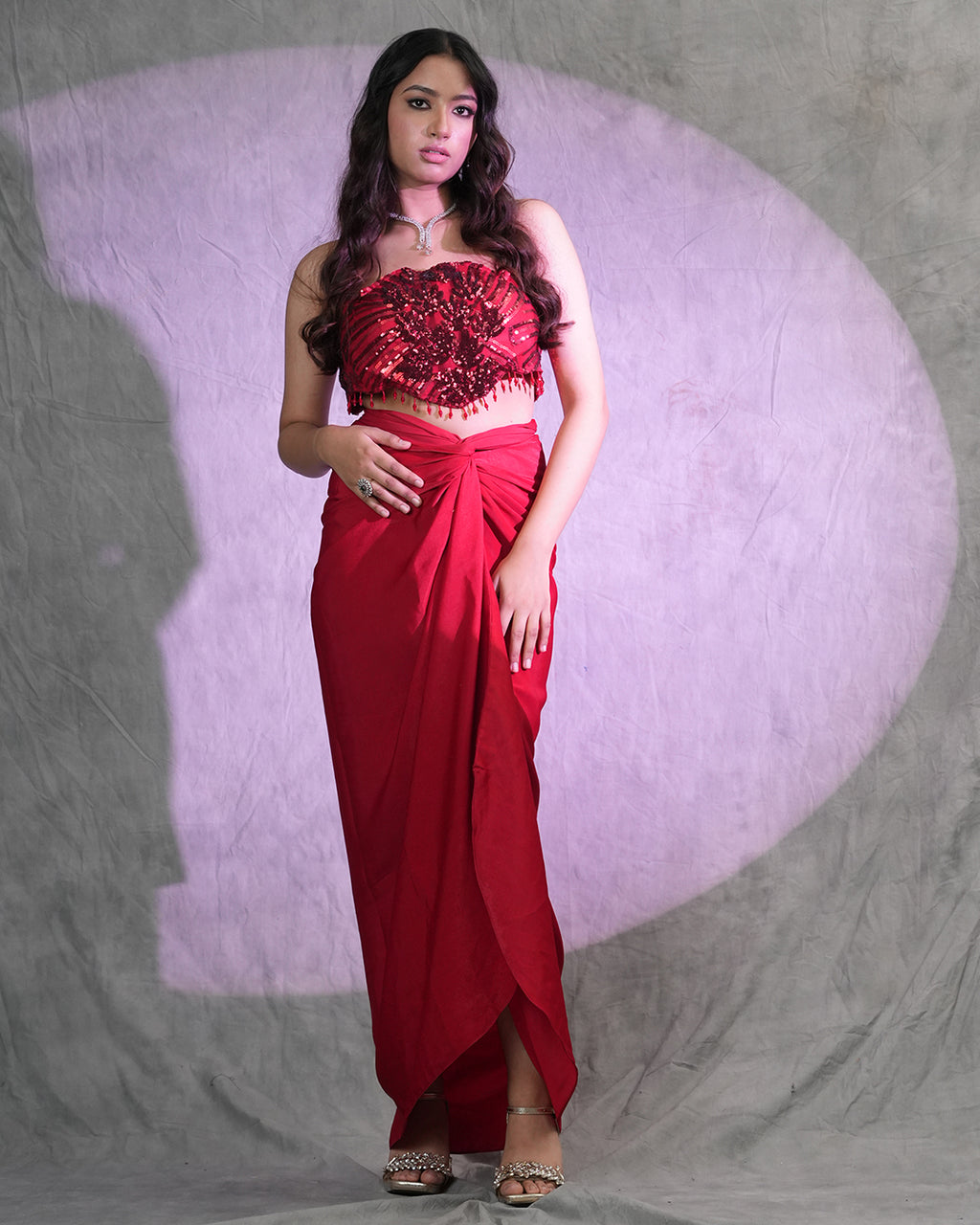 A woman modeling a red two-piece traditional outfit consisting of a heavily embellished red crop top and a matching red draped skirt, standing against a textured gray backdrop with a purple spotlight effect.