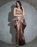 A woman models a metallic bronze and rose gold with a strapless bodice, peplum waist, and draped skirt, set against a textured gray backdrop with a spotlight effect.