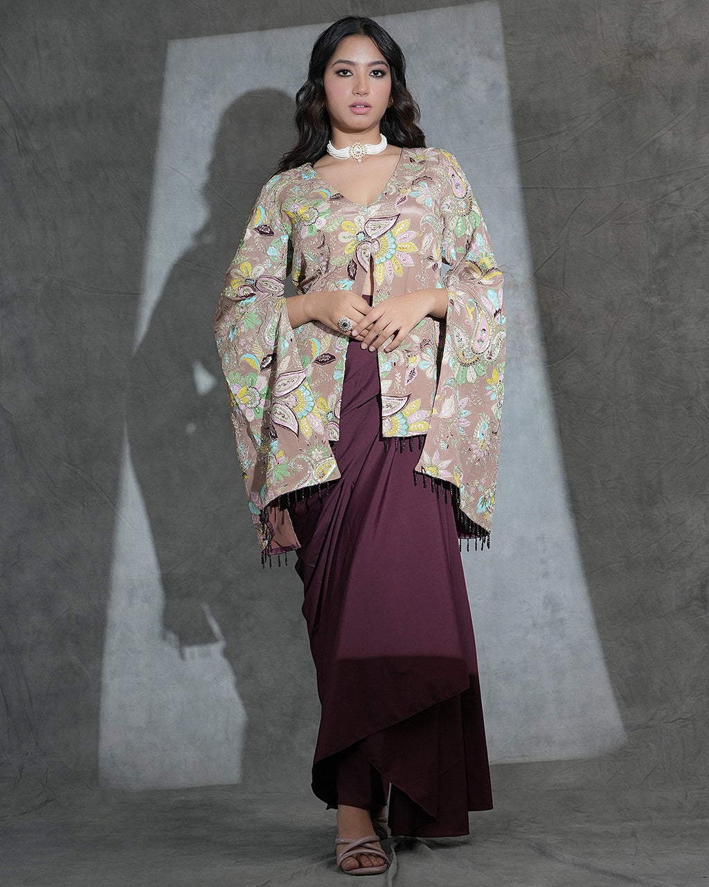 A woman models a traditional outfit featuring a beige and multicolor floral printed shawl draped over a deep maroon dress, set against a textured gray studio backdrop with dramatic lighting.