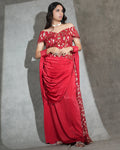 A woman modeling a red off-the-shoulder saree with gold geometric patterns and a matching red dupatta, standing against a textured gray studio backdrop.