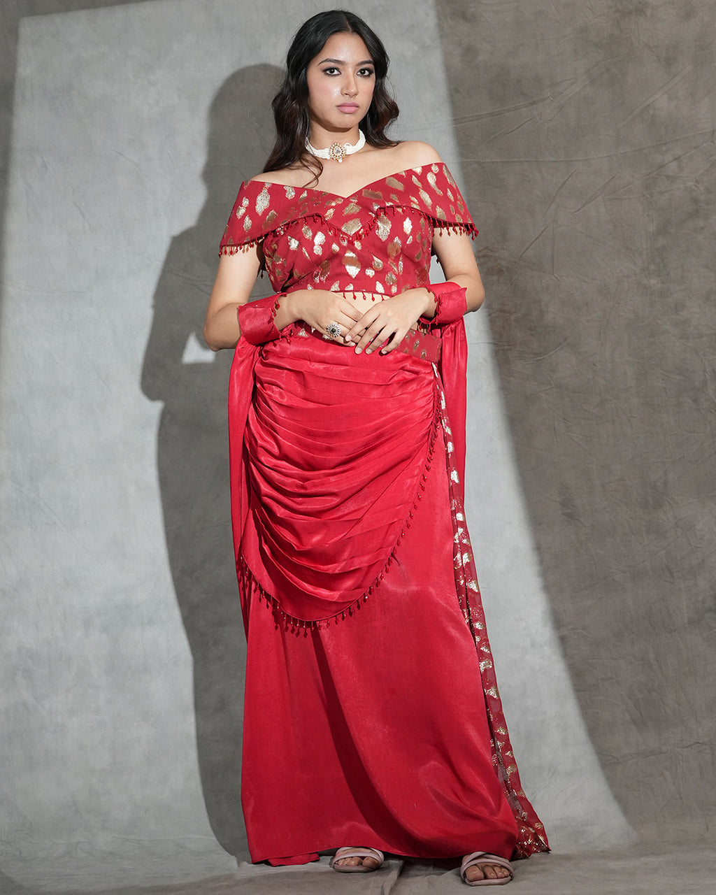 A woman modeling a red off-the-shoulder gown with gold floral embroidery on the blouse and border, standing against a textured gray backdrop.
