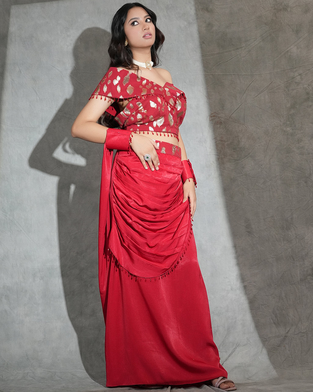 A woman models a red off-the-shoulder saree with gold and white floral patterns on the blouse and a matching draped skirt, posed against a textured gray backdrop with a strong shadow.