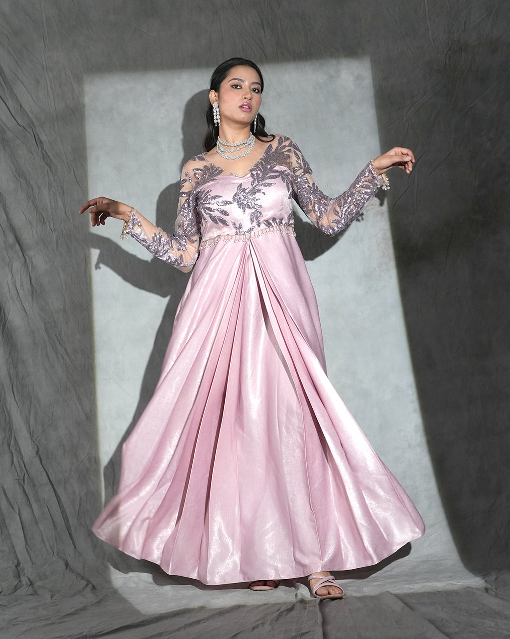 Formal pink gown with sheer floral lace sleeves and a pleated skirt, posed against a gray studio backdrop with dramatic lighting.