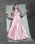 Formal pink gown with sheer floral lace sleeves and a pleated skirt, posed against a gray studio backdrop with dramatic lighting.