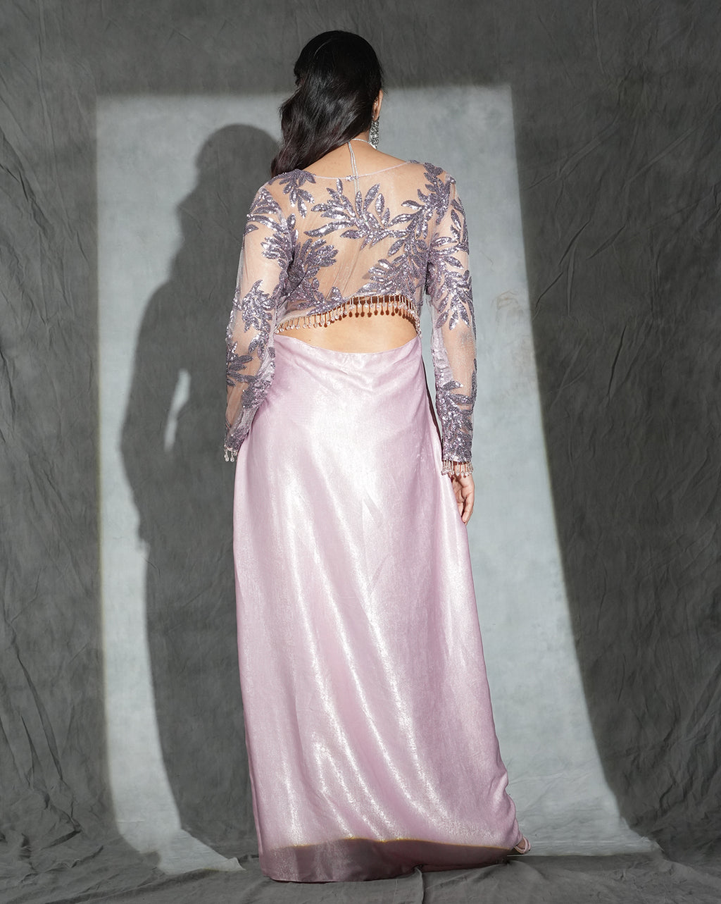A woman shown from the back wearing a floor-length formal gown with a shimmering pale pink skirt and a sheer embroidered top in lavender-purple tones, posed against a textured gray backdrop with a spotlight behind her.